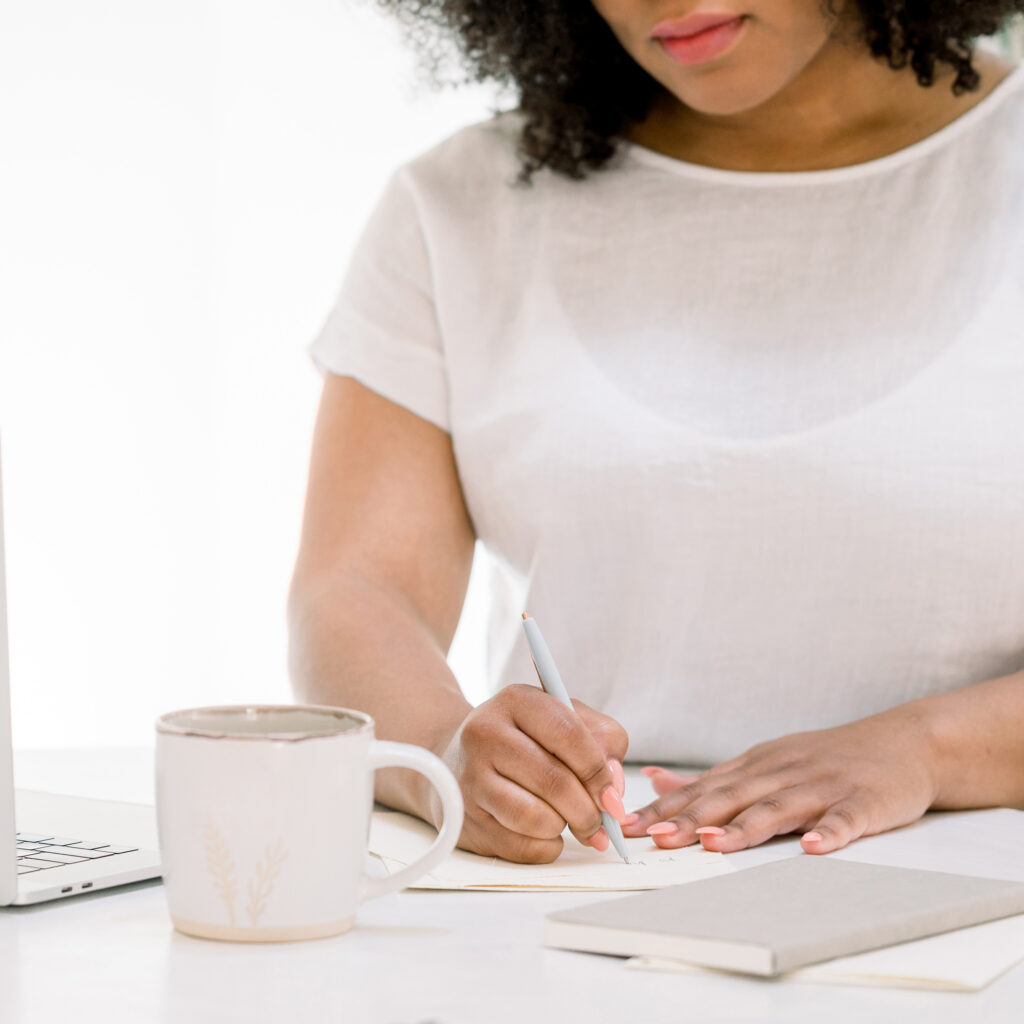 A woman writes in a notebook at a desk with a coffee mug, laptop, and closed book in front of her.