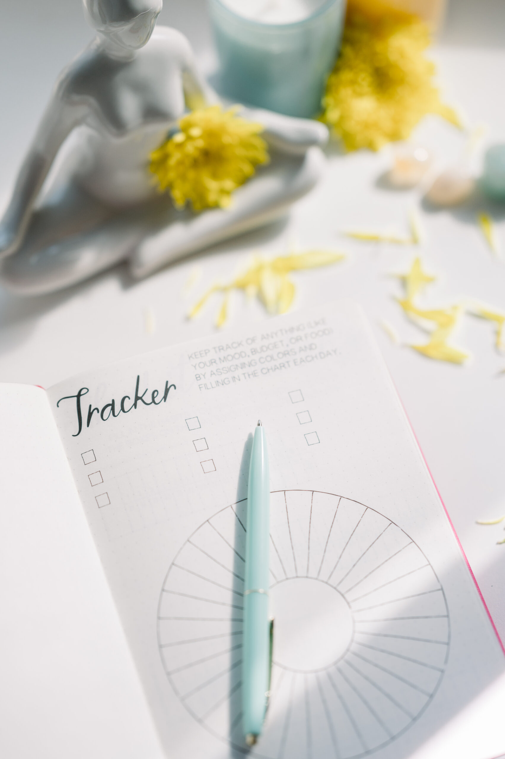Open journal with a tracker chart and pen on the page, beside a gray statue, yellow flowers, and scattered flower petals.